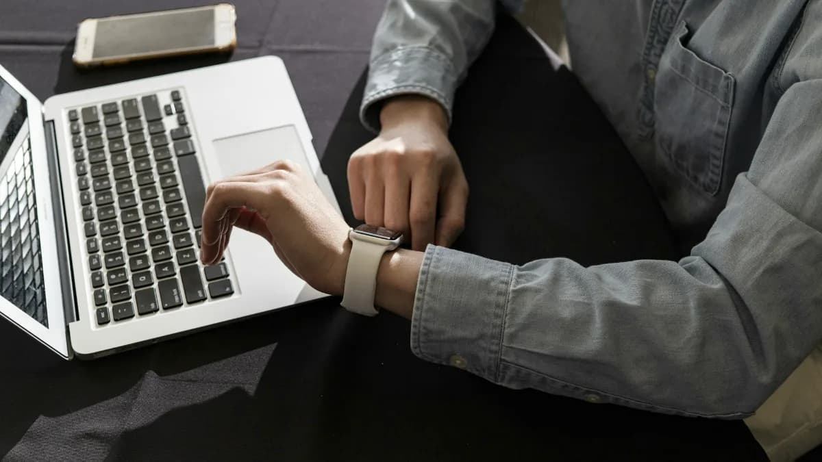 Person sitzt vor Laptop und schaut auf Armbanduhr
